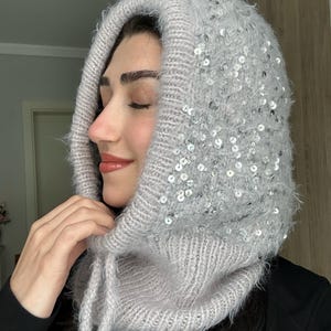 May include: A light gray knitted balaclava hood featuring a fuzzy, textured yarn finish and scattered silver sequins throughout the fabric. The headpiece includes a ribbed trim around the face opening and two long, matching knitted ties at the neck.