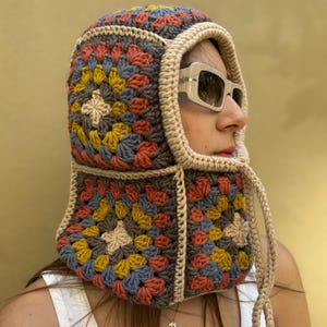 May include: A colorful crochet hood featuring a granny square design in shades of gray, yellow, blue, and orange. The hood has a beige trim and drawstrings. The person is wearing sunglasses and a white tank top.