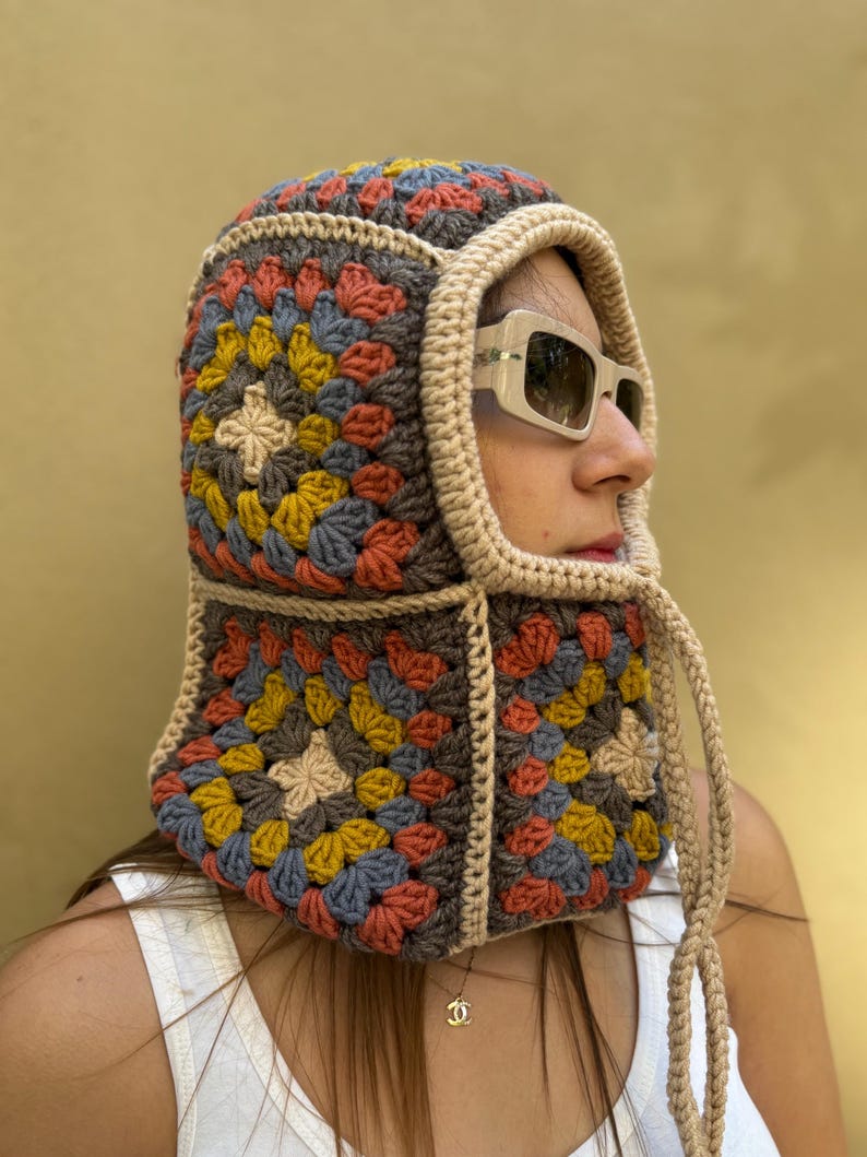May include: A colorful crochet hood featuring a granny square design in shades of gray, yellow, blue, and orange. The hood has a beige trim and drawstrings. The person is wearing sunglasses and a white tank top.