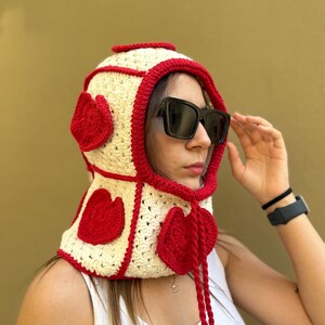 May include: A cream-colored crochet hood with red trim and heart-shaped accents. The hood features a drawstring and is worn by a person wearing sunglasses. The background is a solid olive green.