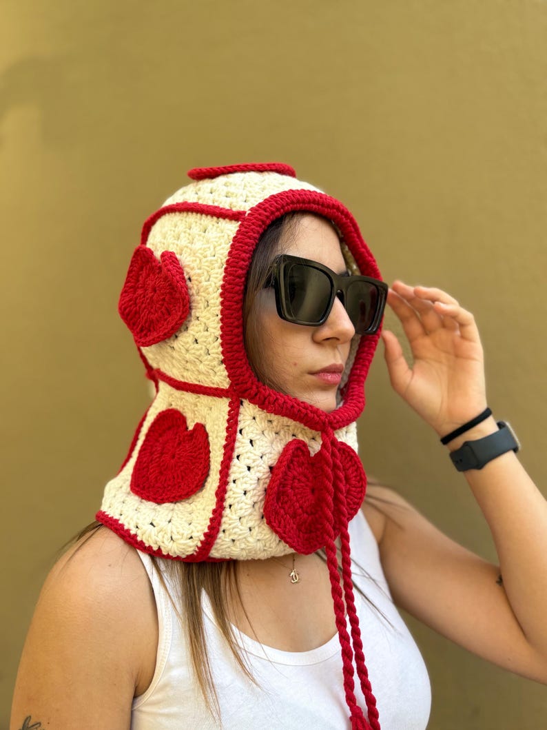 May include: A cream-colored crochet hood with red trim and heart-shaped accents. The hood features a drawstring and is worn by a person wearing sunglasses. The background is a solid olive green.