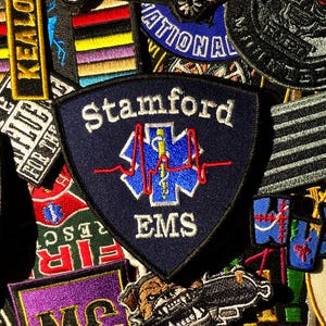 Puede incluir: Una colección de parches bordados, incluyendo un parche azul marino en forma de escudo con el texto "Stamford EMS" y un símbolo médico. Otros parches presentan texto, rayas y varios diseños en una variedad de colores.
