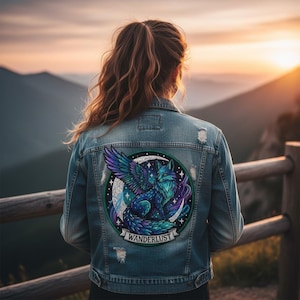 Puede incluir: Chaqueta vaquera de lavado claro con diseño desgastado. La parte trasera presenta un parche circular con un diseño de zorro alado en tonos azules, morados y verdes, con la palabra "WANDERLUST" en una pancarta.