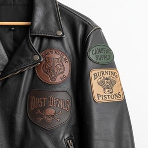 May include: Black leather jacket with various patches. Patches feature a wolf's head, the words "Campfire Supply", "Burning Pistons", and a skull and crossbones with "Dust Devils". The jacket has a zip and snap fastenings.