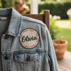 Puede incluir: Chaqueta vaquera azul claro con un parche redondo con el nombre "Olivia" en verde. El parche está rodeado por una corona floral con flores blancas y hojas verdes. La chaqueta tiene detalles clásicos de mezclilla.