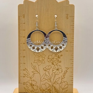 May include: Silver hoop earrings with a geometric design. The earrings are displayed on a wooden stand with a ruler marked in inches. The earrings have a diameter of approximately 2 inches.