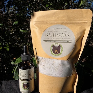 May include: A brown glass bottle of hair oil and a tan paper bag of bath soak. The bath soak bag has a circular label with a bear illustration and the text "BATH SOAK". The hair oil bottle also has a bear illustration and the text "HAIR OIL".