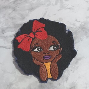 Op de afbeelding: Een geborduurde patch met een gestileerde illustratie van een persoon met een donkere huid, volumineus zwart haar en een felrode strik. De figuur heeft grote ogen met lange wimpers, paarse lippen en rust met de kin op de handen in een geel shirt.