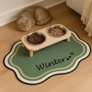 May include: A cat rests near a pet feeding station. The station has two stainless steel bowls on a light wood stand, filled with pet food. The stand sits on a green mat with the word "Winter" and a snowflake design.