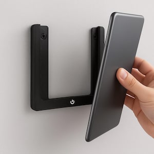 Puede incluir: Un soporte de teléfono negro montado en la pared con un símbolo de encendido. El soporte es un marco en forma de U con un diseño elegante y moderno. Un teléfono inteligente negro se está colocando en el soporte.