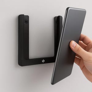 May include: A black wall-mounted phone holder with a power symbol. The holder has a U-shaped frame with a sleek, modern design. A black smartphone is being placed into the holder.