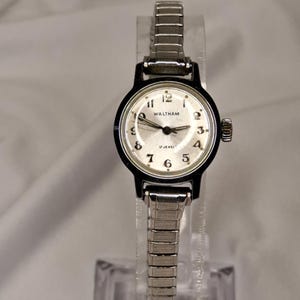 May include: A vintage Waltham wristwatch with a silver face, black bezel, and a silver expansion band. The watch face displays the Waltham logo and the words "17 Jewels". The watch is displayed on a clear plastic stand.