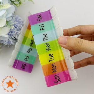 May include: A clear, rectangular weekly pill organizer with seven compartments, each labeled with a day of the week in black text. The compartments are in rainbow colors. The words "GOOD LITTLE GOODS SHOP NAME" are in a circle.