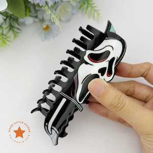 May include: Black and white Scream mask hair claw clip. The hair accessory features a glossy finish and a detailed design of the iconic Ghostface mask. The clip is held by a hand, showcasing its size and shape. The product is from Good Little Goods California USA.