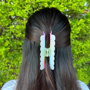 May include: A translucent hair claw clip with a wavy edge design. The clip features a gradient of colors, including pink, blue, and yellow, and is set against dark hair and a green background. The clip is designed to hold hair in place.
