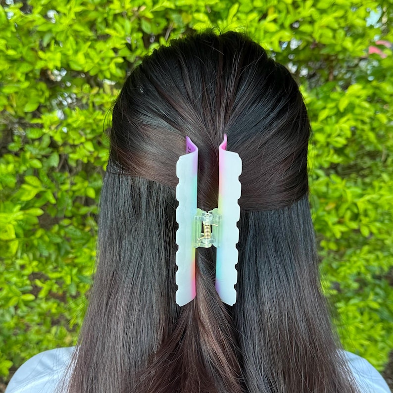 May include: A translucent hair claw clip with a wavy edge design. The clip features a gradient of colors, including pink, blue, and yellow, and is set against dark hair and a green background. The clip is designed to hold hair in place.