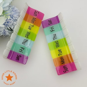 May include: Two rectangular, clear plastic pill organizers with rainbow-colored compartments labeled with days of the week. The text "Sun Mon Tue Wed Thu Fri Sat" is printed on each organizer. The shop name "GOOD LITTLE GOODS" is visible.