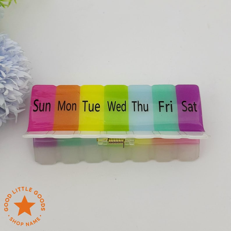 May include: A rainbow-colored weekly pill organizer with compartments labeled for each day of the week. The lid is divided into sections of pink, orange, yellow, green, blue, and purple. The base is translucent with a white hinged lid.