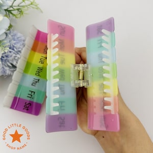 May include: A rainbow-colored pill organizer with compartments for each day of the week. The organizer is held open, revealing the individual compartments. The text "Mon Tue Wed Thu Fri Sat" is visible on the side. The organizer is made of plastic.