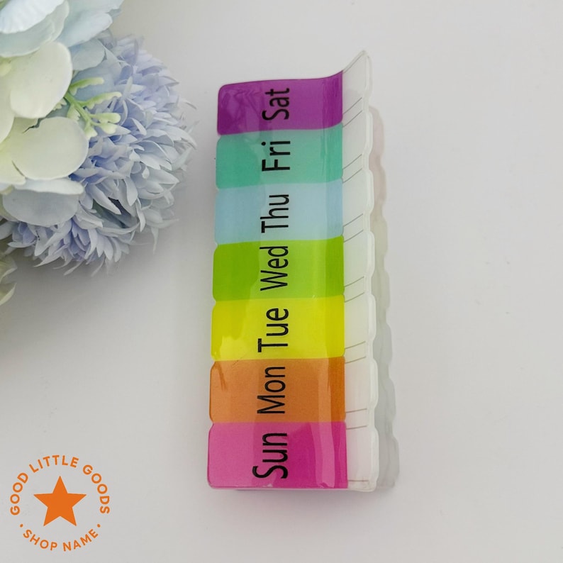 May include: A clear plastic pill organizer with seven compartments, each labeled with a day of the week in black text. The compartments are in a rainbow of colors: pink, orange, yellow, light blue, green, and purple. The product is on a white surface.
