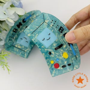 Puede incluir: Dos pinzas para el pelo con estampado en color turquesa y blanco con forma de consola de juegos portátil retro. Cada pinza presenta una pantalla azul claro con una cara sonriente, junto con botones de colores y las letras "BMC" y "B".