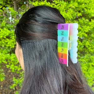 May include: A multi-colored hair clip with the days of the week printed on it. The clip is in the hair of a person with dark hair. The clip is a rainbow of colors, with each day of the week in a different color.