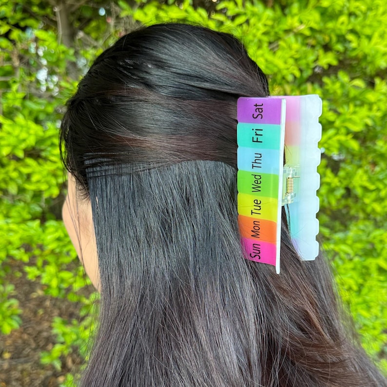 May include: A multi-colored hair clip with the days of the week printed on it. The clip is in the hair of a person with dark hair. The clip is a rainbow of colors, with each day of the week in a different color.