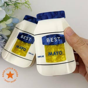 Może przedstawiać: Dwa białe pojemniki w kształcie majonezu z niebieskimi wieczkami i etykietami z napisem "BEST MAYO". Etykiety zawierają również tekst "30FL OZ". Pojemniki mają żółte, teksturowane wnętrze.
