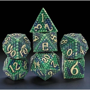 May include: Set of seven polyhedral dice with a dragon scale design in green and blue. The numbers are in a cream color. The dice are for role-playing games like Dungeons and Dragons.