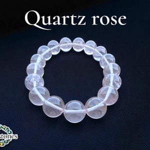 Könnte beinhalten: Ein Armband aus Rosenquarz mit großen, runden Perlen. Die Perlen haben eine durchscheinende rosa Farbe und sind zu einer kreisförmigen Form aufgereiht. Der Text "Quartz rose" wird oben im Bild angezeigt. Das Armband befindet sich vor einem dunklen Hintergrund.