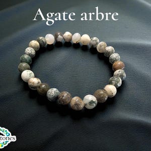 Könnte beinhalten: Ein Armband aus Achatperlen in Weiß-, Braun- und Grüntönen. Das Armband ist kreisförmig und liegt auf einem dunklen, strukturierten Hintergrund. Der Text "Agate arbre" befindet sich oben im Bild.
