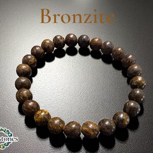 Op de afbeelding: Een Bronzite kralenarmband. De armband heeft ronde, gepolijste kralen in bruine en gouden tinten. Het woord "Bronzite" wordt in bruine letters bovenaan de afbeelding weergegeven. De armband wordt op een zwart oppervlak gepresenteerd.