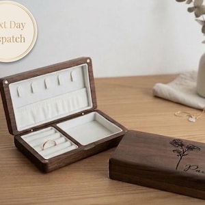 May include: A dark brown wooden jewellery box, open to show ring and earring storage with a light-coloured interior. The closed lid features a floral design and the name "Paula" engraved. The box is on a wooden surface.