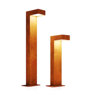 Può includere: Due lampade da esterno rettangolari in stile moderno con una calda finitura marrone ruggine. Le lampade hanno una luce quadrata nella parte superiore e sono montate su una base quadrata. La lampada più alta è alta circa 122 cm, e la più corta circa 76 cm.