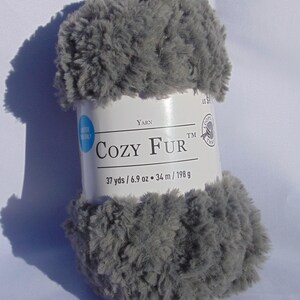 Puede incluir: Ovillo de lana gris Cozy Fur. La etiqueta dice "COZY FUR" e incluye las medidas: 34 m / 198 g. La lana tiene una textura suave y esponjosa, ideal para proyectos de tejido o ganchillo. El ovillo es de color gris claro.