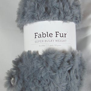 May include: A skein of gray Fable Fur yarn, super bulky weight. The label reads "Koshka 71 yards/100 grams". The yarn has a soft, furry texture, ideal for knitting or crocheting projects. The yarn is a solid gray color.