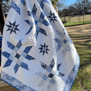 May include: A white and blue quilt with star and geometric patterns. The quilt features various shades of blue against a white background. The border has a floral design. The quilt is draped over a white ladder.