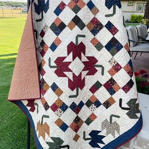 Könnte beinhalten: Eine handgefertigte Steppdecke mit cremefarbenem Hintergrund und einem geometrischen Muster. Die Steppdecke zeigt Quadrate in verschiedenen Farben, darunter Burgunderrot, Blau und Braun, sowie blattartige Formen. Die Steppdecke wird im Freien ausgestellt.