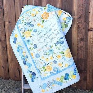 Puede incluir: Un edredón floral con un fondo azul claro, con flores amarillas y azules. El edredón tiene un borde con un diseño de patchwork en tonos azules y verdes. El texto en el edredón dice: "Admire the roses, Drink in the blue skies, Turn your face to the sun, your SPIRIT will fly."