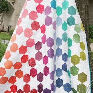 Pode incluir: Um edredom colorido com fundo branco e um padrão de formas hexagonais gradientes. Os hexágonos transitam de laranja para vermelho, rosa, roxo, azul e verde. O edredom está drapeado sobre uma árvore.