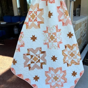 May include: A handmade quilt featuring a white backdrop and a repeating pattern of star-shaped blocks in peach, brown, and floral prints. The quilt is decorated with brown star accents and has a peach-coloured border.