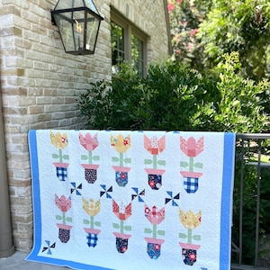 May include: A white quilt with a blue border featuring tulip designs in various colors and patterns. The tulips are in flower pots with different patterns. The quilt is displayed outdoors.
