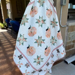 May include: A handmade quilt with a white background, featuring peach-colored pumpkin shapes and green star patterns. The quilt has a red and white striped border and is hanging on a white pole.