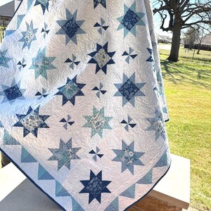 May include: A white quilt with a repeating star pattern in shades of blue and teal. The quilt features a border of blue and teal triangles. The stars have intricate patterns within them, adding visual interest to the handmade textile.