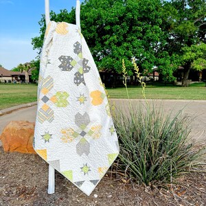 May include: A white quilt with a geometric pattern of gray, yellow, and green shapes, including stars and cross-like designs. The quilt is draped over a white ladder outdoors, with a backdrop of green trees and a blue sky.