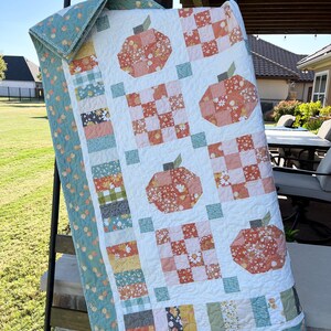 Könnte beinhalten: Eine handgefertigte Steppdecke mit einem Kürbismotiv. Die Steppdecke hat einen weißen Hintergrund mit orangefarbenen und floralen Kürbisapplikationen. Der Rand und die Rückseite sind helltürkis mit einem floralen Muster. Die Steppdecke wird auf einer schwarzen Leiter ausgestellt.