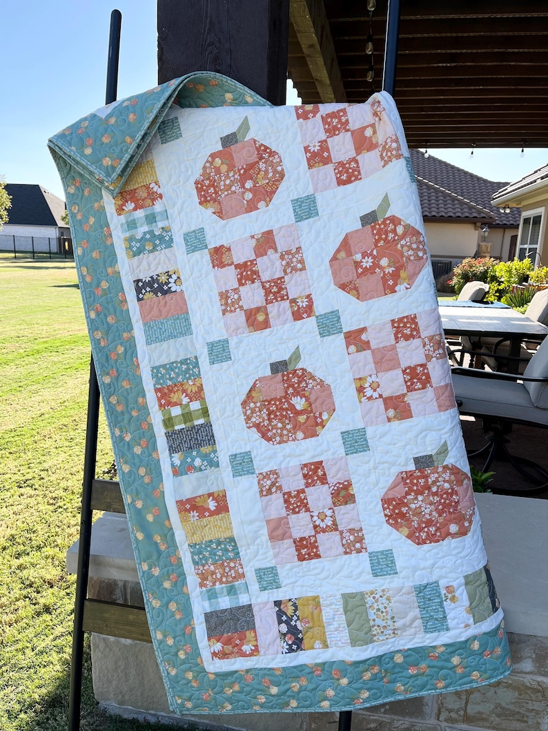 K&ouml;nnte beinhalten: Eine handgefertigte Steppdecke mit einem K&uuml;rbismotiv. Die Steppdecke hat einen wei&szlig;en Hintergrund mit orangefarbenen und floralen K&uuml;rbisapplikationen. Der Rand und die R&uuml;ckseite sind hellt&uuml;rkis mit einem floralen Muster. Die Steppdecke wird auf einer schwarzen Leiter ausgestellt.