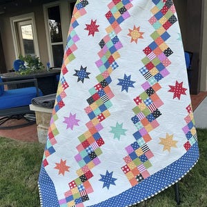 Op de afbeelding: Een kleurrijke quilt met een witte achtergrond, met zigzagpatronen van patchwork vierkanten in verschillende kleuren. De quilt is versierd met stervormen in meerdere kleuren en heeft een blauwe stippenrand. De quilt wordt buiten tentoongesteld.
