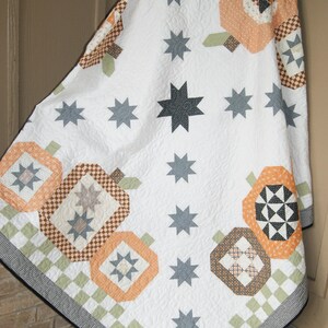 May include: A white quilt with a pumpkin and star pattern. The quilt features orange, brown, and grey pumpkin shapes, along with grey stars. The border has a checkered pattern in green, white, and black.