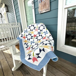 Op de afbeelding: Een handgemaakte quilt met een sterrenpatroon in rood, geel, blauw en groen op een witte achtergrond. De quilt is gedrapeerd over een witte Adirondack-stoel op een houten veranda. De quilt heeft een blauwe stippelrand.